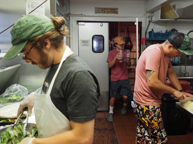 Three people are working in a kitchen; one is mixing a bowl of food, another is preparing ingredients on a counter, and the third is holding a drink near the entrance.