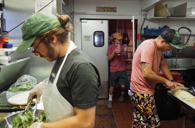 two people work in a kitchen — one mixing a salad and another slicing meat, as a third walks in while carrying cups.