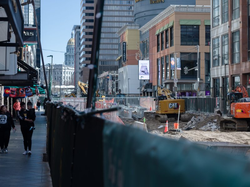 City street under construction with heavy machinery and barriers. People walking on the sidewalk to the left, buildings and a distant dome visible in the background.