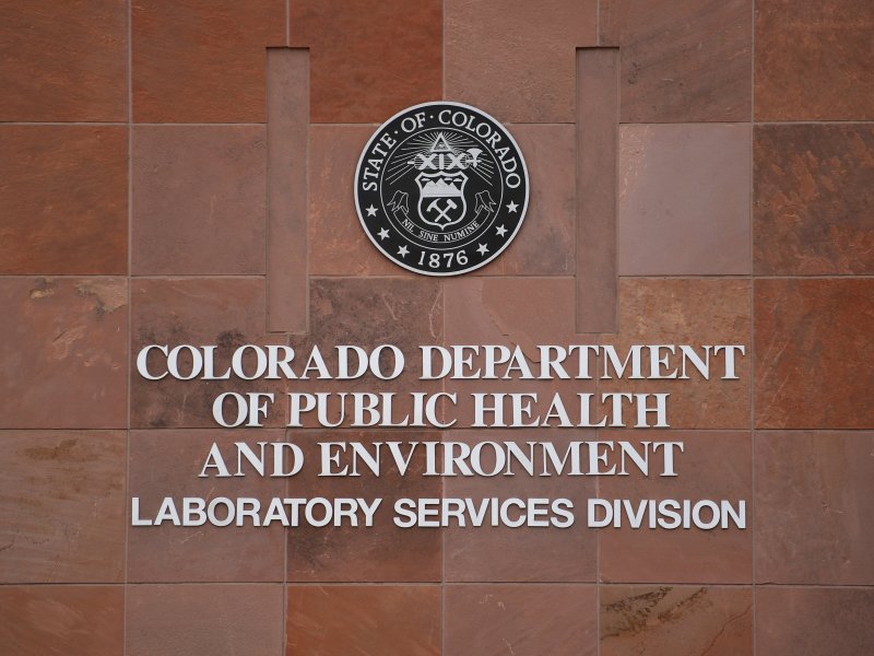 The sign for the Colorado Department of Public Health and Environment's Laboratory Services Division, featuring the state seal, is displayed on a red brick wall.