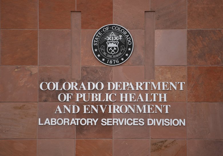 The sign for the Colorado Department of Public Health and Environment's Laboratory Services Division, featuring the state seal, is displayed on a red brick wall.