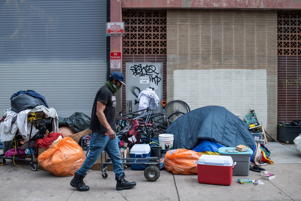 A person walks past a makeshift encampment with a tent, personal belongings, and miscellaneous items on a city sidewalk, highlighting the pressing issue of homelessness.