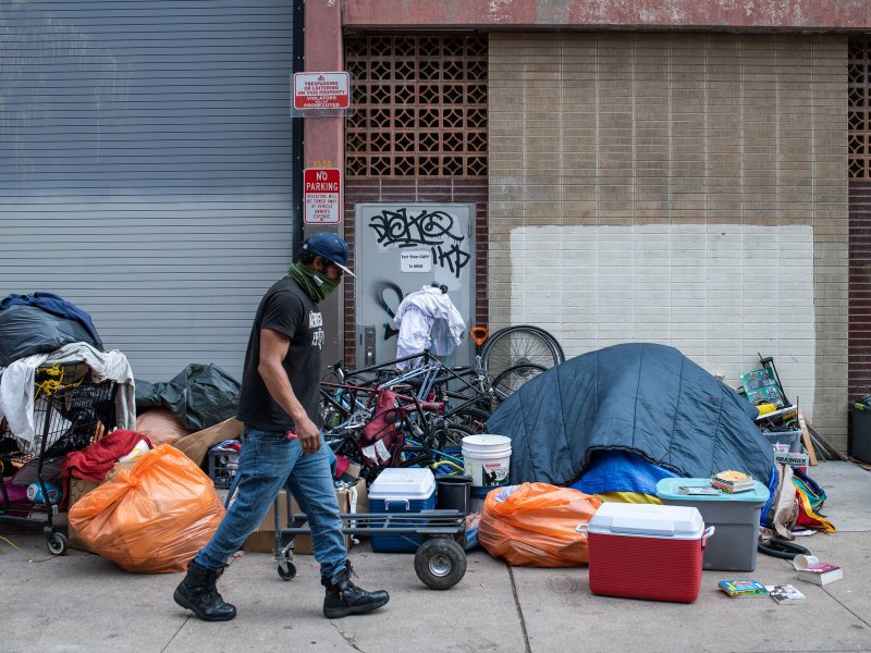 A person walks past a makeshift encampment with a tent, personal belongings, and miscellaneous items on a city sidewalk, highlighting the pressing issue of homelessness.