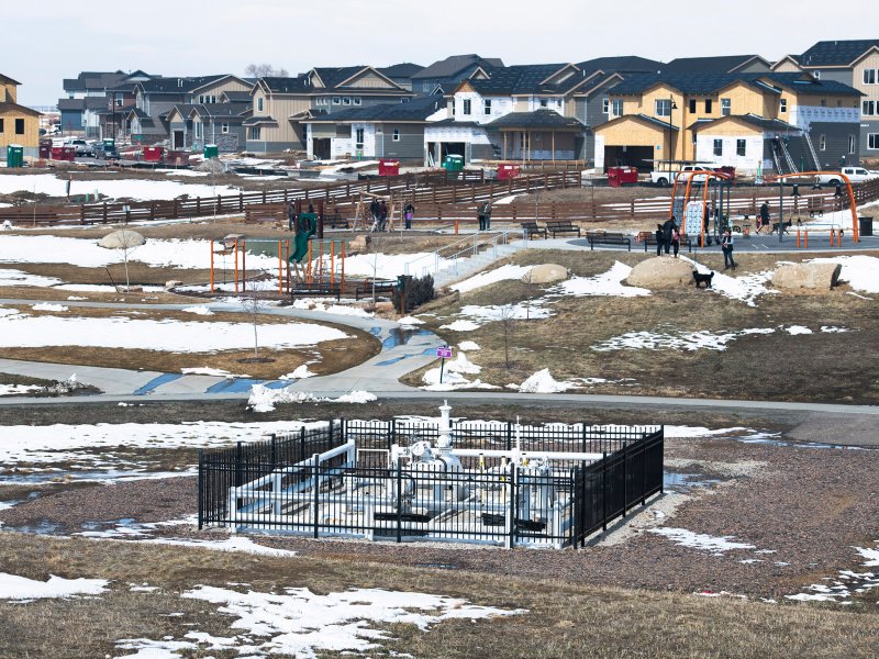 A suburban neighborhood showcases snow patches on the ground, scattered houses, a fenced-in utility area, and a playground with people in the distance. A cloudy sky overhead adds to the sense of air pollution.