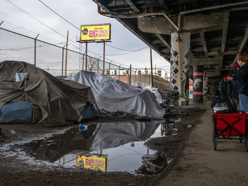 A person with a red cart stands near a row of makeshift tents under an overpass, highlighting the stark reality of homelessness. A large puddle on the ground nearby reflects a billboard for an energy drink in the background.