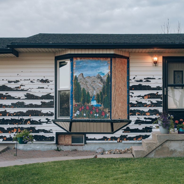 A house with peeling paint and a bay window featuring a mural of mountains and trees. Grass and stairs with potted plants are in the foreground.