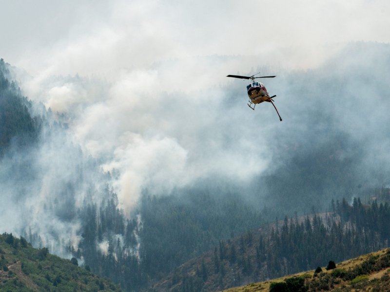 A helicopter flies above a forested area engulfed in smoke, presumably responding to a wildfire on a mountainside.