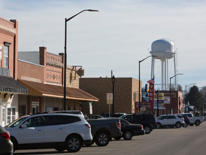 A small-town street is lined with parked cars and brick buildings. A tall water tower is visible in the background. The street is quiet under a partly cloudy sky.