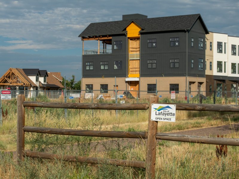 Newly constructed buildings stand behind a wooden fence with a "City of Lafayette Open Space" sign, surrounded by a dry grassy area.