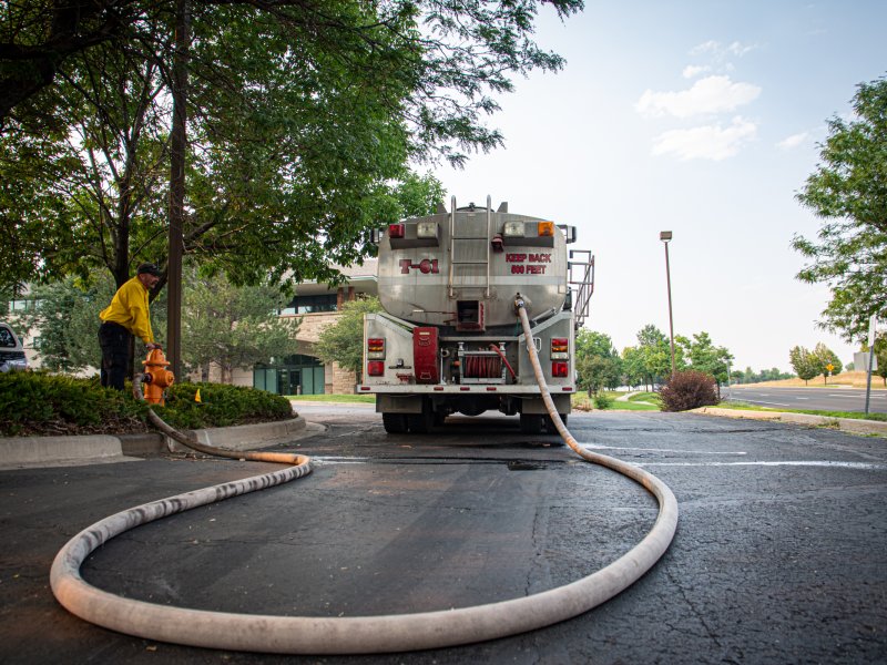 Worker filling a tanker truck with water from a fire hydrant on a tree-lined street.