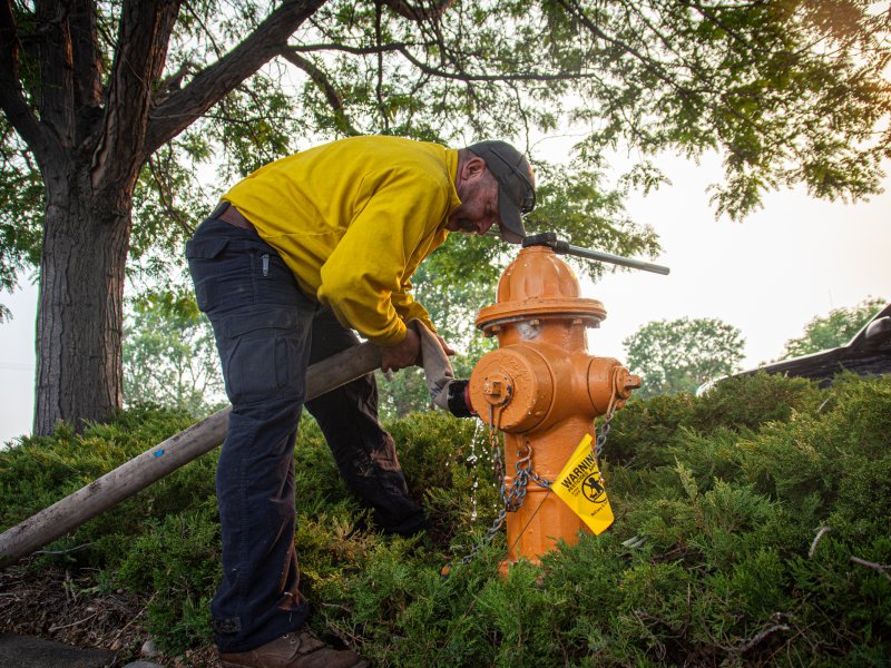 A man in a yellow shirt and black cap uses a tool to work on an orange fire hydrant in a green, landscaped area.