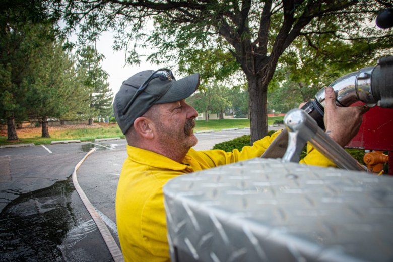 A firefighter in a yellow uniform attaches a hose to equipment on a fire truck outdoors, with trees and a parking lot in the background.