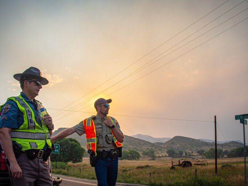 Two uniformed officers stand on a roadside with a sunset in the background, next to a black police vehicle. The landscape features hills and open fields under a partly cloudy sky.