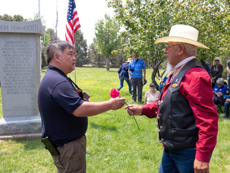 A man hands a flower to another man in front of a stone monument.