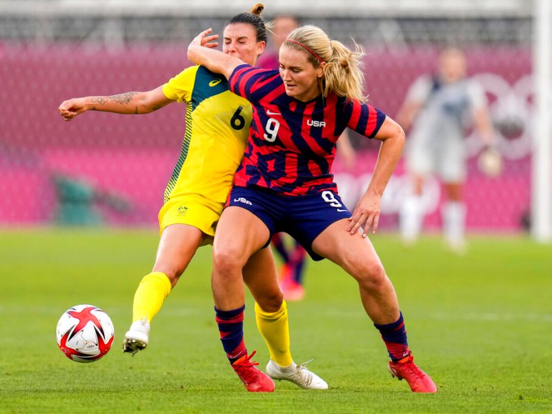 Two soccer players — Lindsey Horan and Chloe Logarzo — in action during a match. The player in yellow attempts to tackle the player in red and blue, who is holding the ball. The goalkeeper stands in the background near the goal.