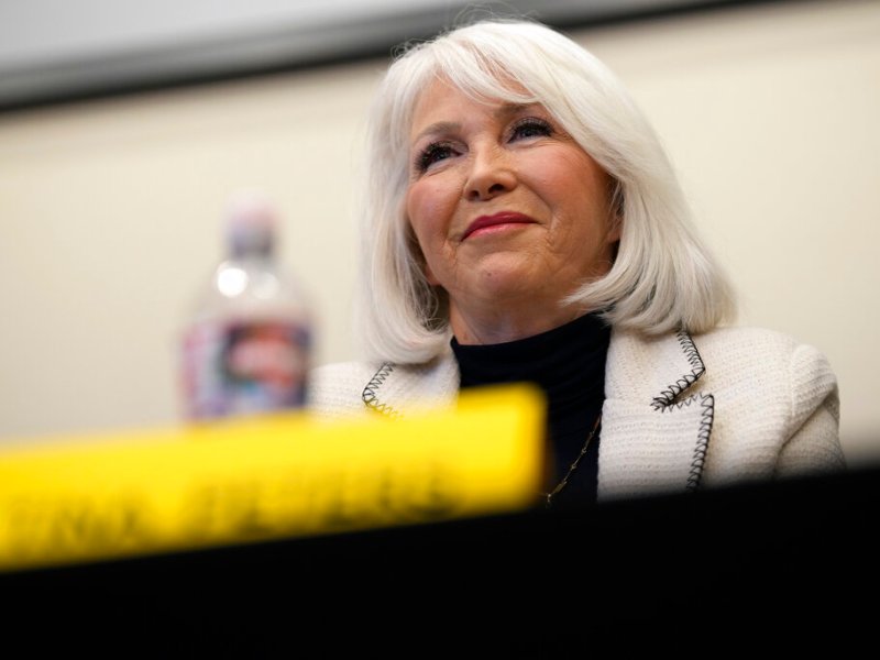 Tina Peters, with white hair in a white jacket and black turtleneck, sits at a table, with a yellow name placard and a plastic water bottle in front of her.