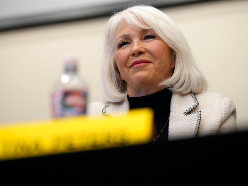 Tina Peters, who has white hair, is seated, wearing a white jacket and black top, with a water bottle and a yellow name tag in front of her.