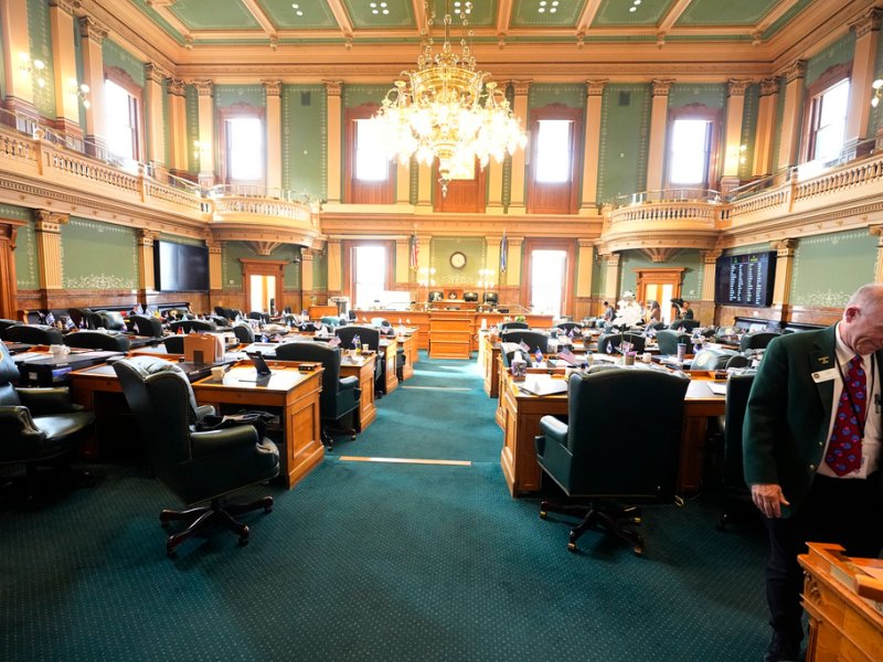 A legislative chamber with green carpet and wooden desks; a chandelier hangs from the ornate ceiling. Members in business attire converse and move around the room.