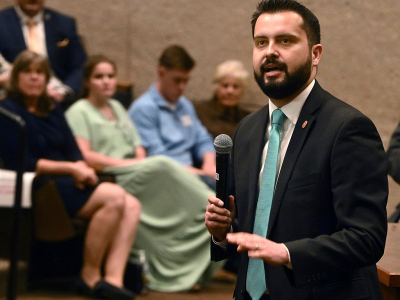 Dave Williams, wearing a black suit and blue tie, talks into a microphone. People sit in a crowd listening.