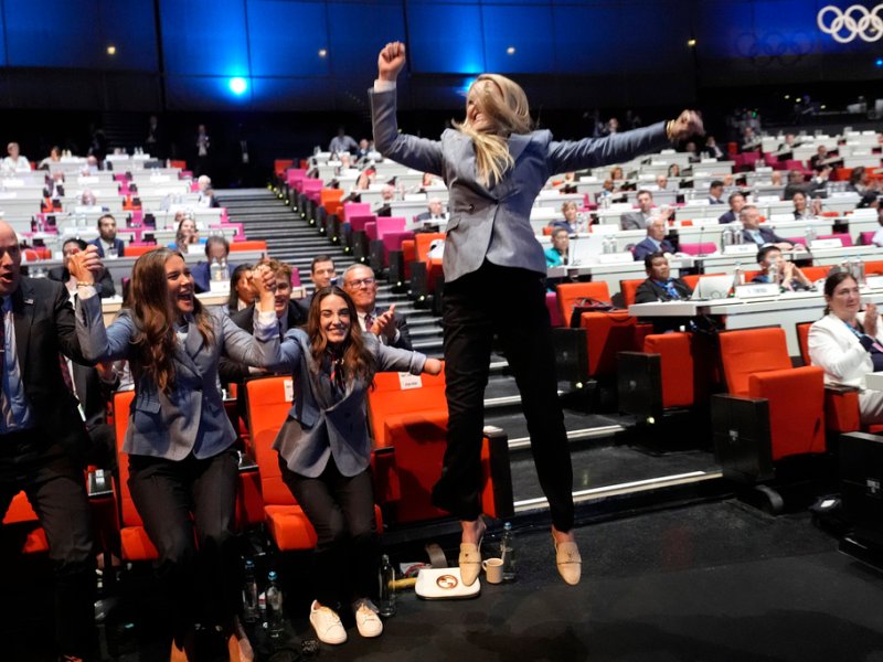 People in business attire cheer and celebrate at an event held in a large auditorium with an Olympic logo in the background.