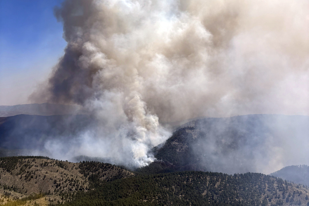 A large plume of smoke rises from a wildfire burning through a forested mountain area under a clear sky.