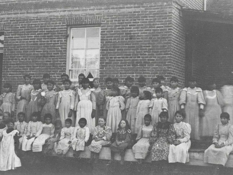 A historical black-and-white photo of a group of Native American children in dresses, posing in front of a brick building.