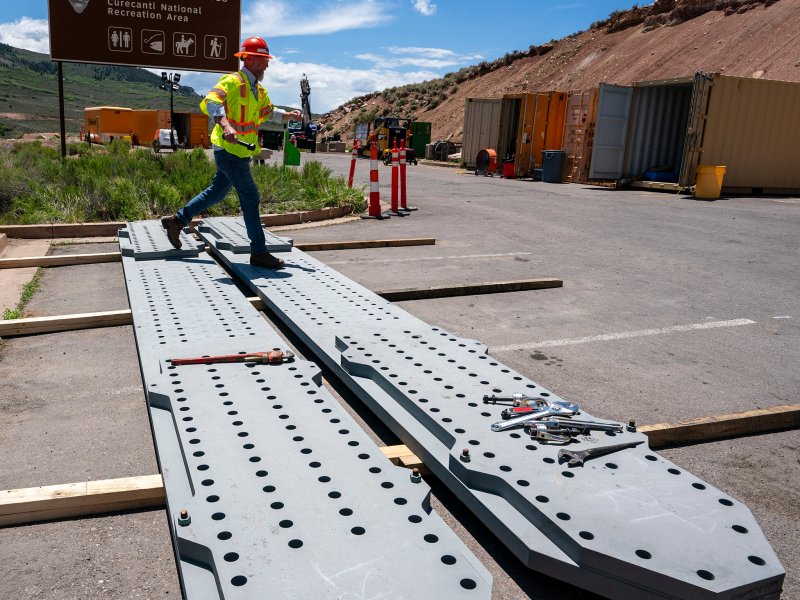 A construction worker in a hard hat and safety vest walks past large metal beams and tools at Dillon Pinnacles, Curecanti National Recreation Area.