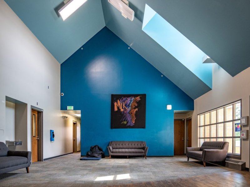 A spacious room with high ceilings and skylights. There are three gray couches, a blue accent wall with a colorful abstract painting, and wooden doors leading to other rooms.