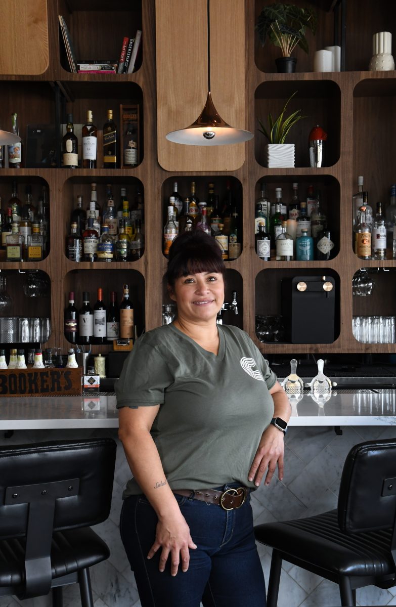 Dana Rodriguez stands in front of a bar stocked with various bottles of alcohol. She is wearing a green t-shirt and jeans and is smiling at the camera.