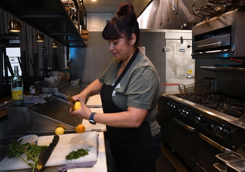 A person in a grey t-shirt and black apron is peeling a vegetable on a cutting board in a commercial kitchen. Various kitchen utensils and appliances are visible in the background.