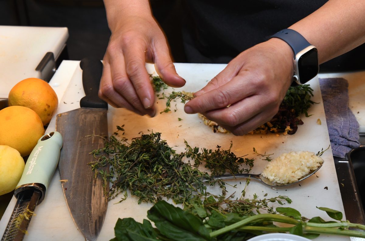 Dana Rodriguez is preparing food, seasoning with herbs on a countertop. A large kitchen knife and a spoonful of minced garlic are visible on the cutting board amidst various ingredients.