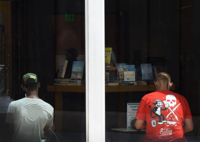 Two individuals lean against the window of a library. A shelf of books is seen in the background. 