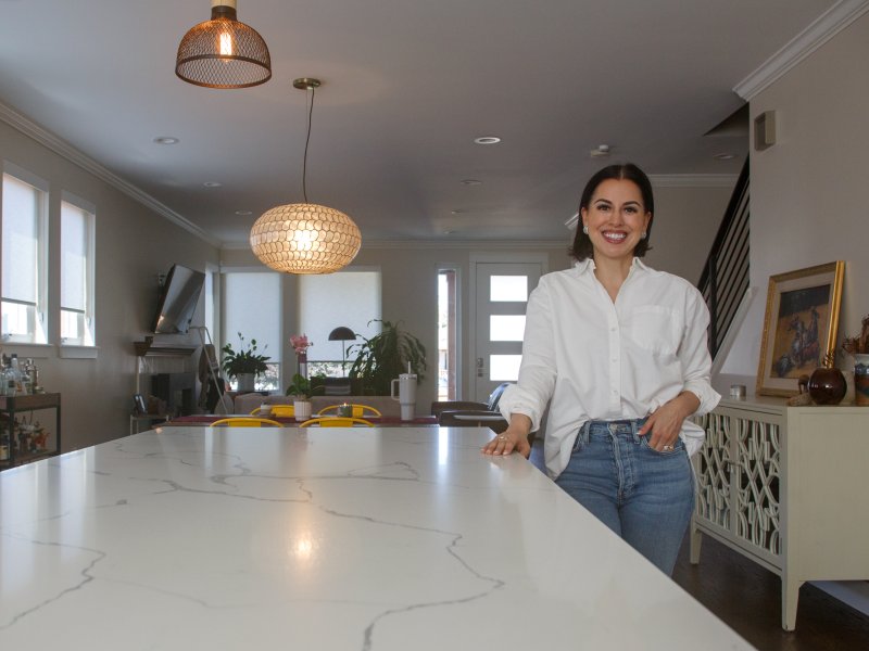 A woman in a white shirt and jeans stands by a marble kitchen island in a modern, well-lit kitchen-dining area.