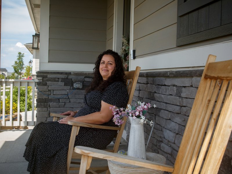 A person with long, curly hair sits on a wooden rocking chair on a porch. There's another empty wooden rocking chair beside them. A small table with a metal vase holding flowers is nearby.