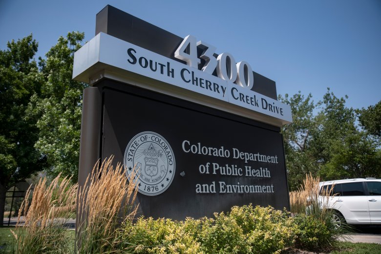 Sign displaying "4300 South Cherry Creek Drive" and "Colorado Department of Public Health and Environment" with the state seal of Colorado, surrounded by vegetation and a parked car nearby.