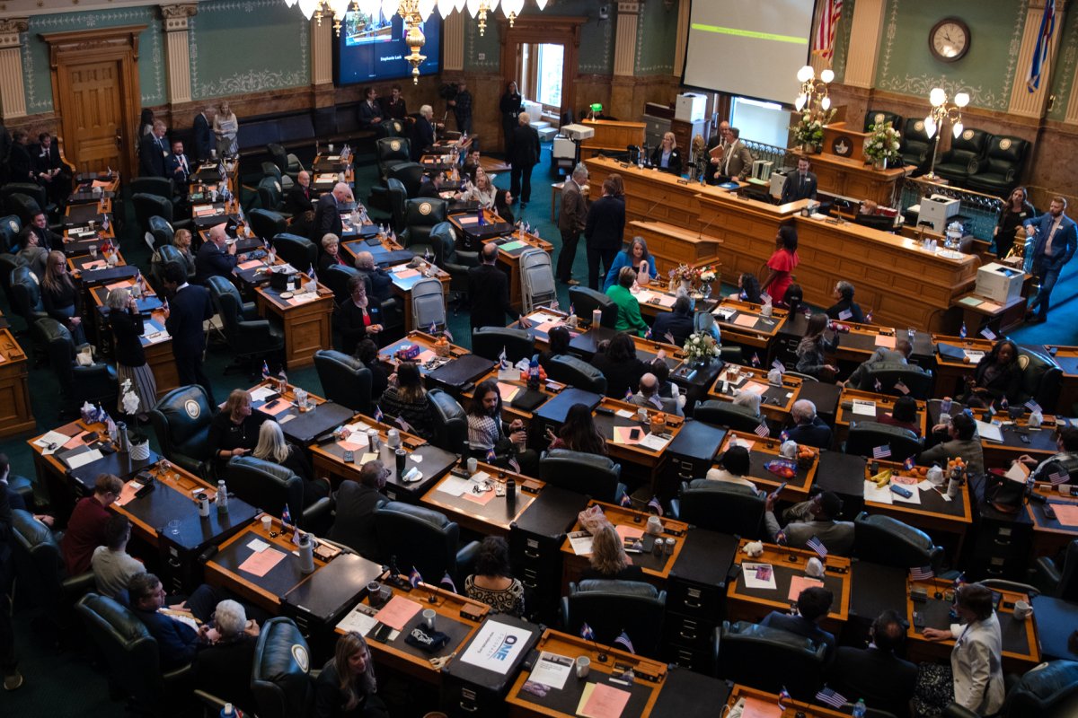 A legislative chamber filled with people seated at desks and standing, engaging in discussions and activities, with a few officials seated at a central elevated desk.