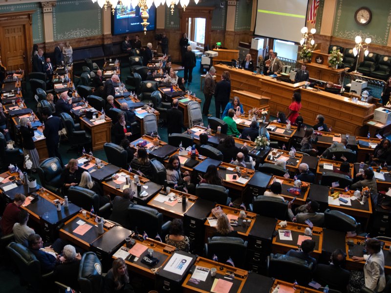 A legislative chamber filled with people seated at desks and standing, engaging in discussions and activities, with a few officials seated at a central elevated desk.