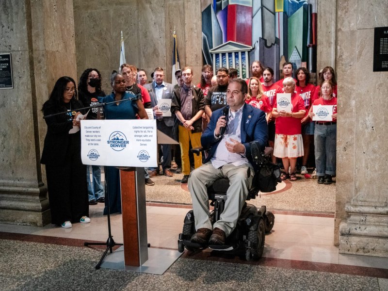 A group of people facing the camera, with one person speaking at a podium and another in a wheelchair holding a microphone. The backdrop includes a colorful mural and marble walls.