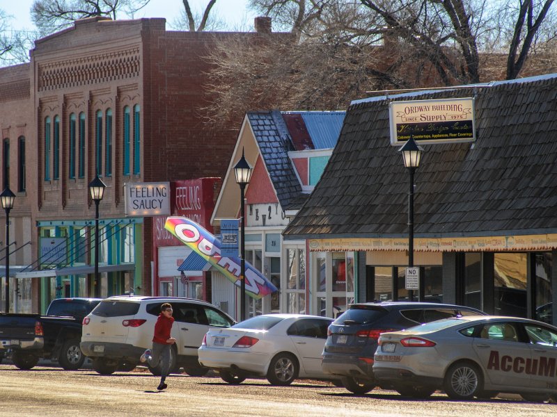 A row of parked cars in front of various shops, including one with a colorful sign, on a sunny day with some people walking on the sidewalk.