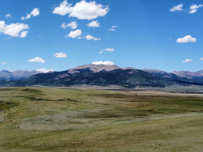 A wide, open plain with green fields under a blue sky with scattered white clouds. Mountains are visible in the background.