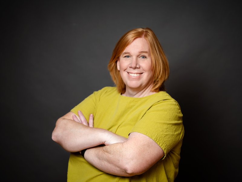 A person with short red hair, wearing a yellow top, smiles with arms crossed against a dark background.