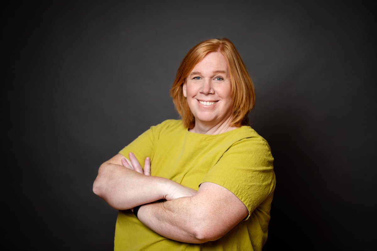 A person with short red hair, wearing a yellow top, smiles with arms crossed against a dark background.