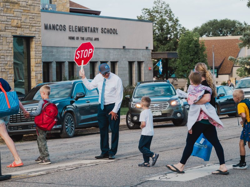 A crossing guard holds a stop sign, assisting children and parents across a crosswalk in front of Mancos Elementary School.