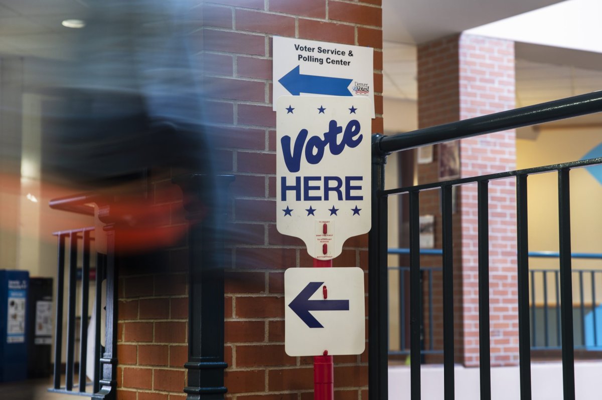 A "vote here" sign with an arrow pointing left hangs on the outside of a building with a blurred figure walking past.