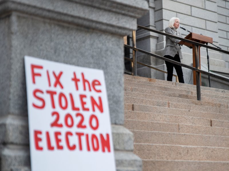 Tina Peters speaks at a podium on steps next to a large sign that reads, "Fix the STOLEN 2020 ELECTION.