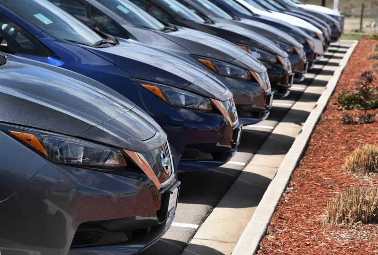 A row of parked cars is lined up in a parking lot, with their front ends facing the camera. The cars are mostly silver and blue, and are neatly aligned.