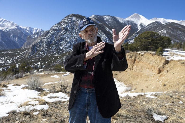 An older man with blue baseball camp and dark suit gesturing with his hands as he talks outside with snowy peaks in the background.