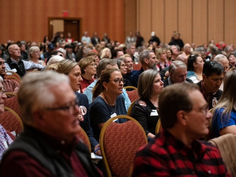 A large group of seated people attentively listen during a conference or seminar in a room with orange walls and wood paneling.