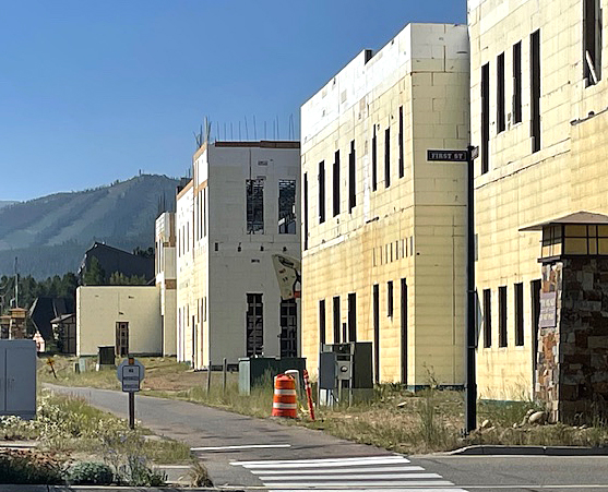 A construction site with several unfinished buildings, some wrapped in insulation material. An orange traffic cone and a "First St" street sign are visible on a sunny day.