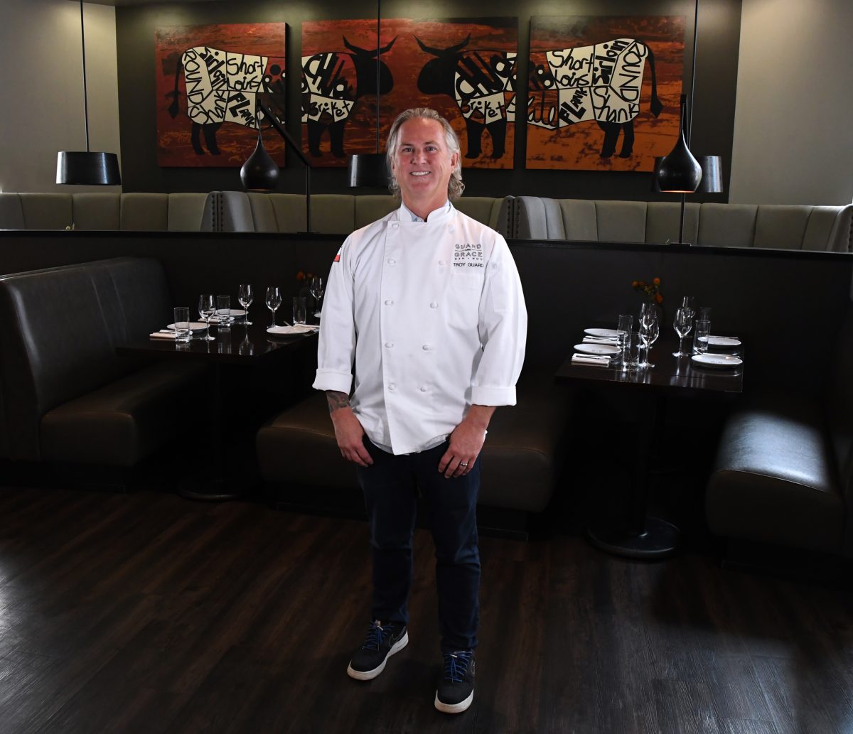 Troy Guard, a chef wearing a white coat and dark jeans, stands in a restaurant dining area with modern decor, featuring leather booth seating and abstract art on the walls.